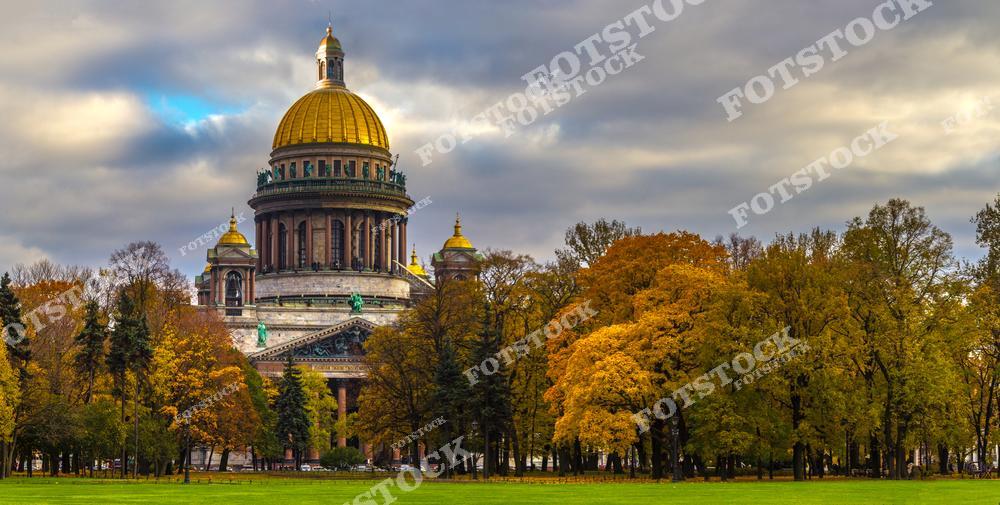 На снимке запечатлен Исаакиевский собор — один из самых величественных храмов Санкт-Петербурга. Золотой купол собора ярко выделяется на фоне облачного осеннего неба. Деревья перед собором покрыты разноцветной листвой, что придает пейзажу теплые осенние оттенки. Этот вид Исаакиевского собора осенью подчеркивает гармонию архитектуры и природы в центре города.